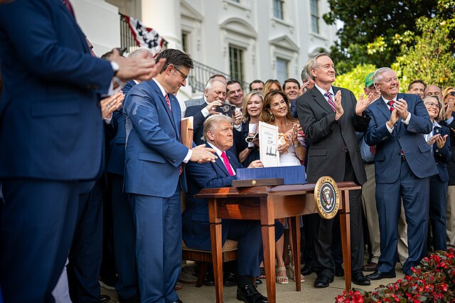 President Donald Trump signs the One Big Beautiful Bill Act outdoors on the South Lawn of the White House, surrounded by applauding officials and supporters. The scene captures a formal bill signing ceremony with a large group present, symbolizing leadership and legislative achievement.